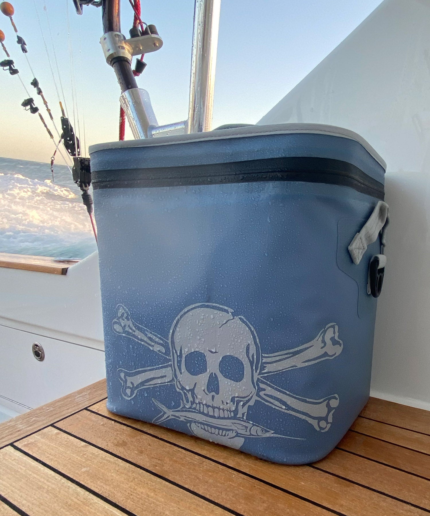 A blue Calcutta cooler with a white skull and crossbones graphic sits on a wooden deck on a boat, with the ocean and sky visible in the background.